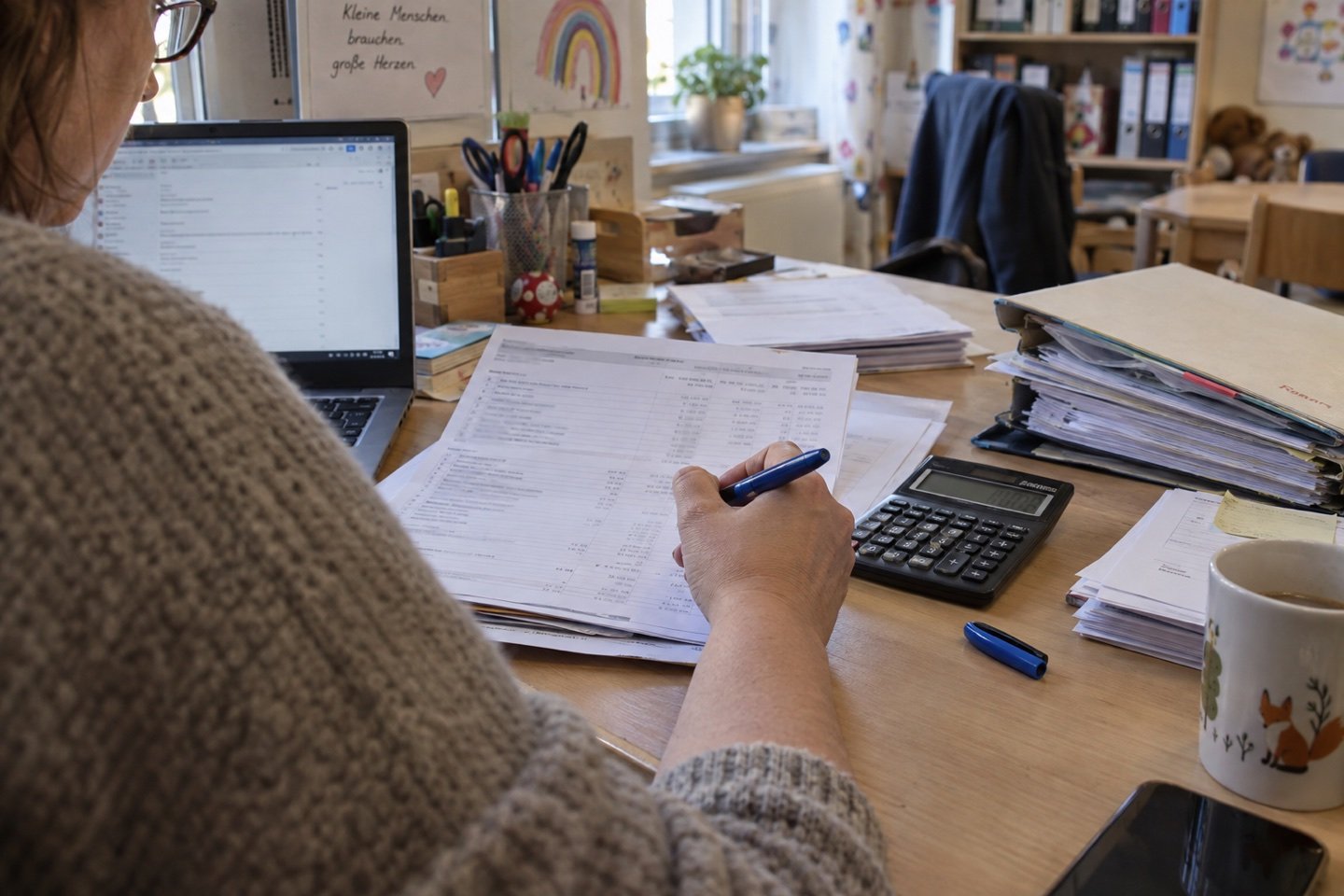 Eine Mitarbeiterin prüft Abrechnungsunterlagen mit Stift und Taschenrechner am Schreibtisch